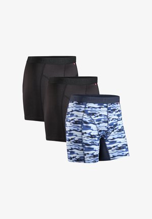 Uomo sorridente senza maglietta indossa boxer a fantasia mimetica blu e grigia, in piedi contro uno sfondo neutro.