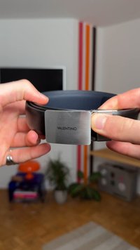 Fekete bőr öv, amelyen egy téglalap alakú ezüst fém csat található, amin a "VALENTINO" felirat látható. Sima textúra, minimalista dizájn és masszív kivitel jellemzi.