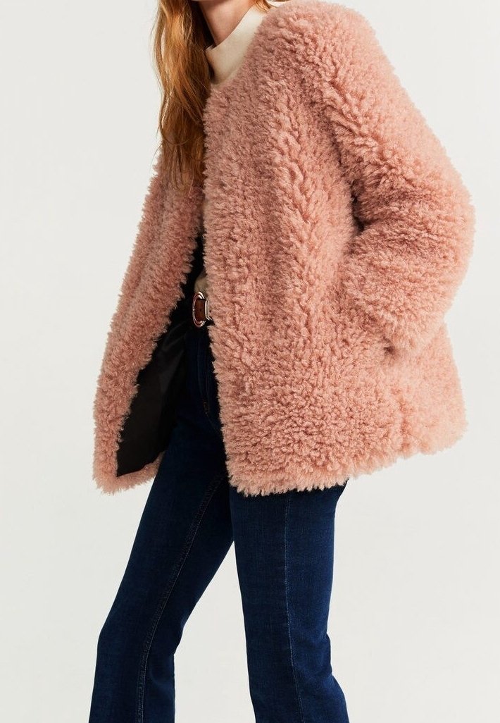 Femme portant un manteau teddy rose duveteux, un col roulé crème, un jean bleu foncé et une ceinture marron, debout avec les mains dans les poches du manteau.