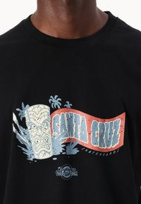 Zwarte katoenen t-shirt met een grafisch ontwerp van een tiki-masker met blauwe en rode accenten, vergezeld van de tekst "SANTA CRUZ" in het vet.