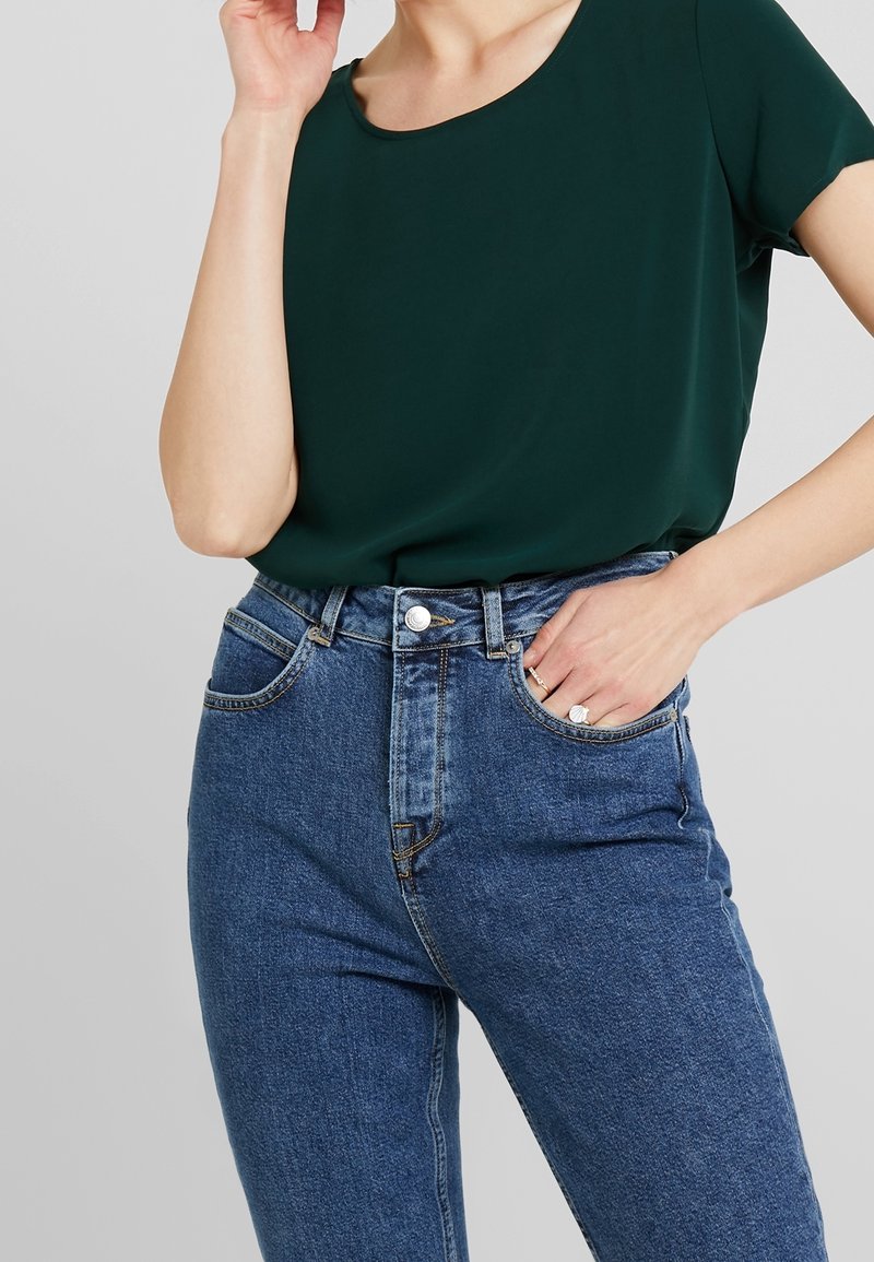 Haut vert foncé à manches courtes associé à un jean en denim bleu taille haute. Le jean présente des poches classiques et une fermeture par bouton.