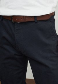 Pantalones de algodón azul marino con una textura suave, complementados por un cinturón de cuero marrón que presenta una hebilla metálica plateada.