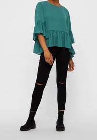 Top teal a maniche corte con rouches a strati, abbinato a jeans skinny neri con strappi all'altezza del ginocchio e stivali neri alla caviglia.