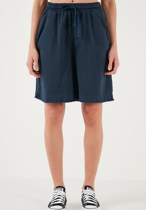 Osoba nosząca granatowe szorty na sznurek z postrzępionymi brzegami oraz czarne niskie trampki Converse, stojąca na jednolitym tle.