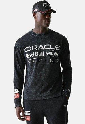 Mann mit schwarzem Langarmshirt und Mütze mit den Logos von "Oracle Red Bull Racing", steht mit einer Hand in der Hosentasche und schaut nach rechts.