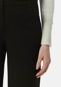 Zwarte op maat gemaakte broek met een gladde textuur, met zijzakken. Een licht geribbelde trui met knoopaccenten is gedeeltelijk zichtbaar bij de mouw.