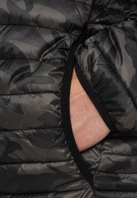 Polstret jakke i sort og mørkegrønt camouflage-mønster. Har vandret quiltemønster og sort kant omkring ærmegabet.