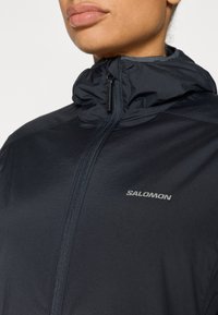 Casaco preto leve com gola alta, fecho de correr frontal e detalhe refletor. Apresenta um logótipo Salomon no peito. Textura lisa.