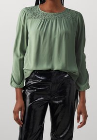 Grön blus med en spetspanel vid halsen, plisserad fram, och långa puffärmar, i kombination med svarta glänsande byxor i ormskinnsmönster.