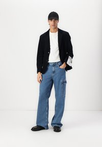 Férfi öltözet fekete blézerrel, fehér pólóval, oversized kék farmertáskával, oldalzsebbel, és fekete cipővel. Fekete sapka a fején.