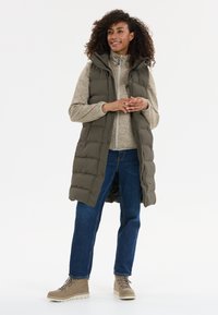 Langer, olivgrüner Puffer-Weste mit Kapuze, über einem hellbeigen Reißverschluss-Fleece, kombiniert mit blauen Jeans und beigen Wanderstiefeln mit Schnürsenkeln.