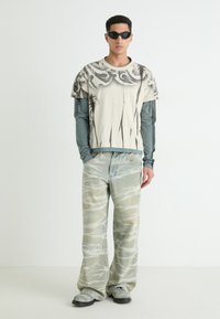 T-shirt graphique blanche superposée sur un haut à manches longues teal, assortie de jeans baggy délavés avec des motifs abstraits et de chaussures grises.