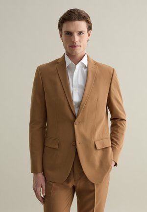 Abito marrone su misura realizzato in tessuto liscio, caratterizzato da una chiusura a due bottoni, rever a punta e tasche laterali, abbinato a una camicia bianca.