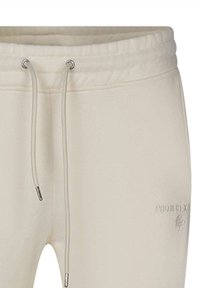 Pantalon de survêtement beige clair avec une taille élastique, fermeture à cordon et poche latérale. Logo "PROJECT X" embossé sur la jambe gauche.