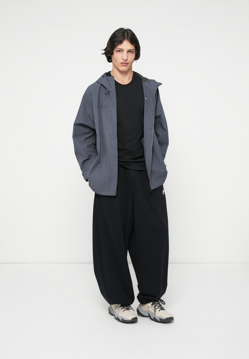 Grijze lichtgewicht jas met capuchon, zwart T-shirt, losse zwarte broek en beige sportschoenen met donkere zolen, staand op een effen achtergrond.