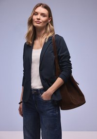 Femme blonde en blazer sombre et jean, main dans la poche, portant un grand sac à bandoulière en suède marron sur un fond uni.