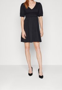 Svart klänning med V-ringning, puffärmar, smockad byst och utsvängd kjol. Bärs med svarta högklackade skor mot en vit bakgrund.