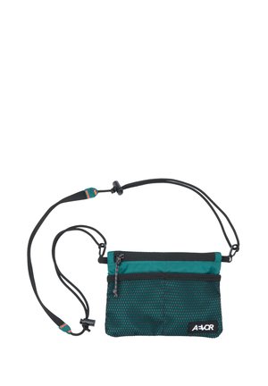 Kompakte Umhängetasche in Türkis und Schwarz mit Netzvordertasche, Reißverschluss, verstellbarem Riemen und AEVOR-Logo in der unteren rechten Ecke.