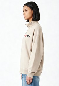Pull-over en polaire beige avec un col haut, une fermeture éclair et un texte brodé en noir et rouge "I ❤️ YOU." Texture douce et coupe décontractée.