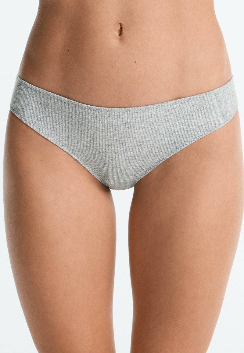 Personne portant une culotte de style bikini côtelé gris clair, montrée de la taille au haut des cuisses sur un fond uni.