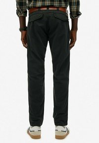 Mand iført sorte bukser, brun bælte, ternet skjorte og hvide sneakers, stående med ryggen mod en ensfarvet hvid baggrund.