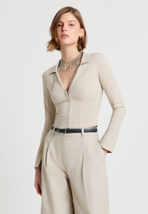 Donna con top beige aderente a maniche lunghe e pantaloni a gamba larga a vita alta con cintura nera, che indossa una collana argentata a frange, in posa con la mano in tasca.