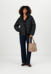 Vrouw met krullend haar, gekleed in een zwarte pufferjack, donkere brede broek, bruine loafers en met een beige suède totebag in haar hand.