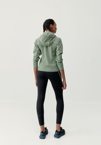 Svetlo olivovo zelená bunda s kapucňou, reflexné detaily a dlhé rukávy, doplnená čiernymi legínami a tmavými teniskami. Minimalistický dizajn.