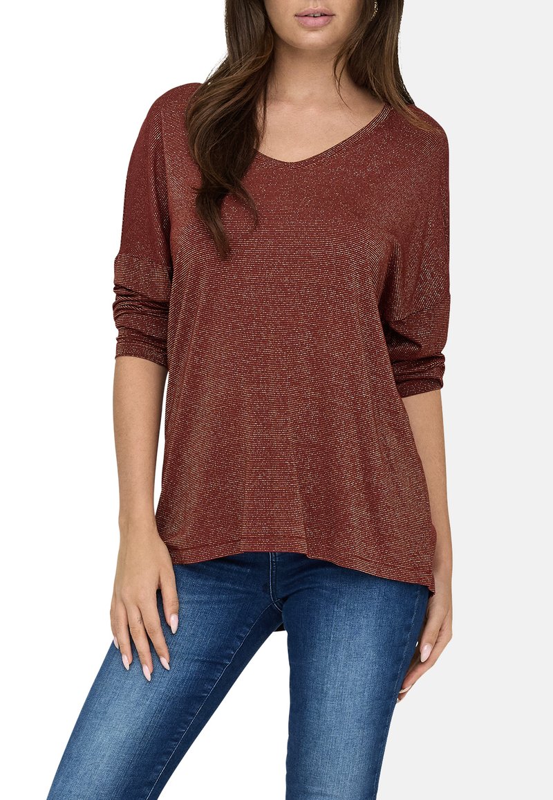 Top oversized de color óxido con escote en V, textura brillante y mangas largas arremangadas en los puños, combinado con jeans de mezclilla azules.