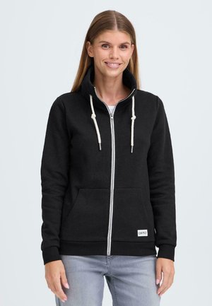 Sort zip-up hoodie med høj krave, kontrast snore, to frontlommer og et hvidt mærke på kanten. Bæres over en hvid skjorte.