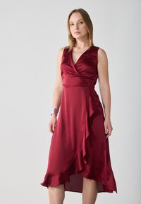 Burgundyröd satinklänning med v-ringning, ärmlös design och asymmetrisk nederkant, med en volangdetalj längs kjolen.