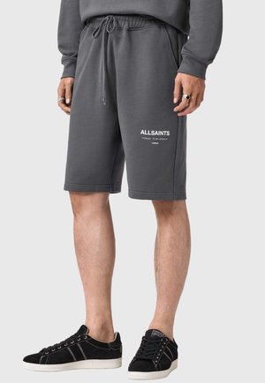 Mann trägt dunkelgraue AllSaints-Shorts mit weißem Logo-Schriftzug, schwarze Sneaker mit weißen Sohlen und Ringe an den Fingern, steht vor einfarbigem Hintergrund.