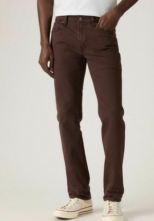 Personne portant un jean slim marron foncé et des baskets blanches basses, debout avec une main dans la poche devant un fond uni.
