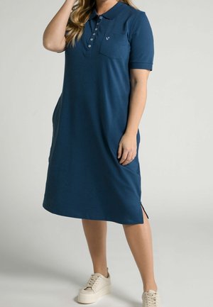 Marineblauwe polokleed van zachte stof, met een kraag, korte mouwen, een borstzakje en zijsplitsen. Gecombineerd met lichte sneakers.