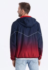 Waterdichte jas die overgaat van marineblauw naar rood, met een capuchon, elastische manchetten en een reflecterend zigzagpatroon op de rug.