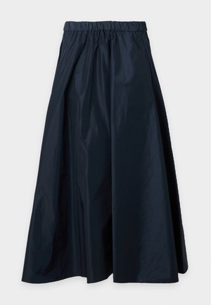 Femme portant un haut rose clair à manches courtes, une longue jupe noire et des chaussures plates noires, debout devant un fond clair uni.