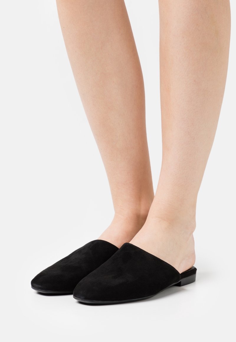 Jambes portant des mules plates noires à enfiler avec des bouts pointus fermés sur fond blanc.