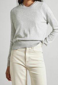 Pull gris clair avec un col et des poignets côtelés, présentant un motif texturé. Associé à un jean beige clair, créant un look décontracté.