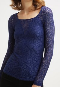 Haut bleu marine à manches longues en dentelle avec des motifs complexes en forme de losange et des dessins tourbillonnants. Présente un col dégagé et une silhouette ajustée.