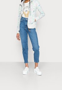 Ljust grå hoodie med blommönster, vit grafisk t-shirt, högmidjade blåjeans och vita platåskor. Avslappnad outfit visas.