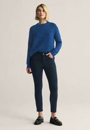 Kvinde med blondt hår iført en blå, blød sweater, mørke, tætsiddende bukser og sorte loafers med guldkædedetaljer, stående foran en ensfarvet baggrund.