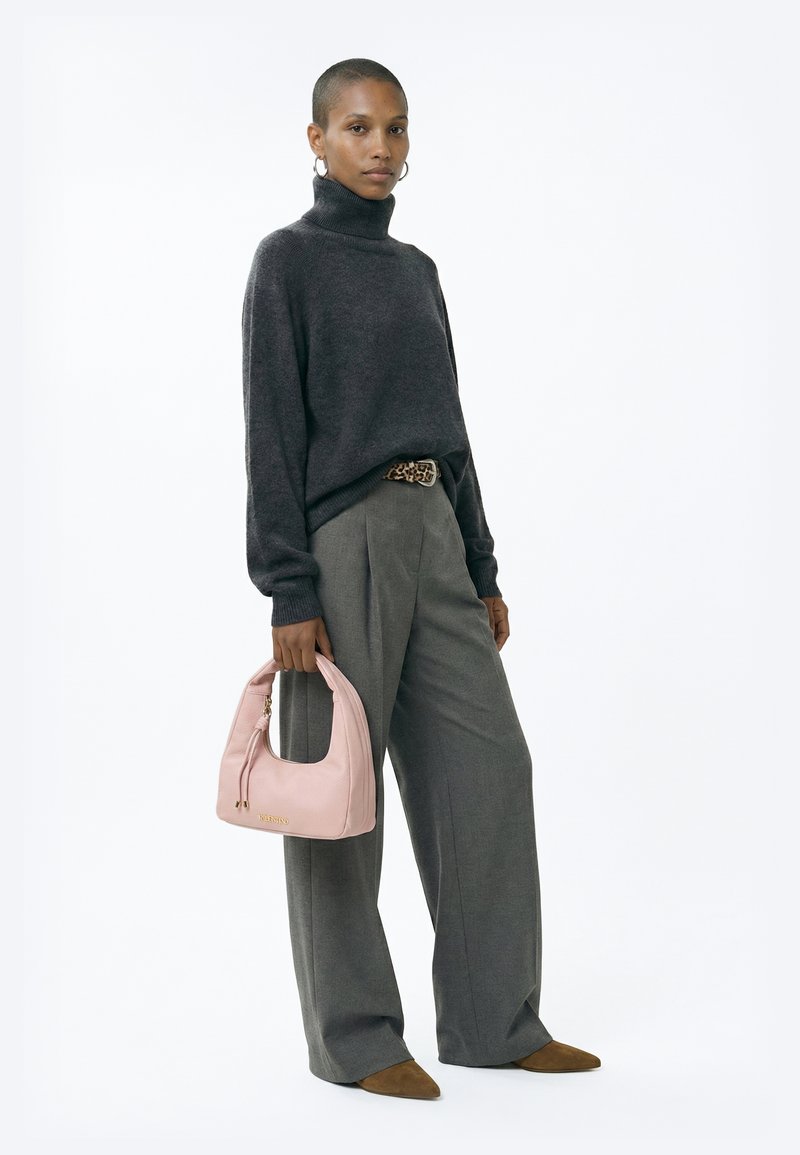 Γυναίκα με σκούρο πουλόβερ με ψηλό γιακά, φαρδιά γκρίζα παντελόνα με ζώνη λεοπάρ, κρατώντας ροζ τσάντα χειρός, στέκεται μπροστά σε λευκό φόντο.