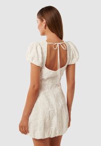 Weißes Kleid mit Puffärmeln, offener Rückseite und einer Schleifenverschluss. Verfügt über strukturierte Blumenmuster auf der gesamten Fläche. Kurze Länge.