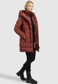 Rust-colored padded coat with a high collar and hood, featuring two side pockets and a streamlined silhouette, paired with black leggings and boots.