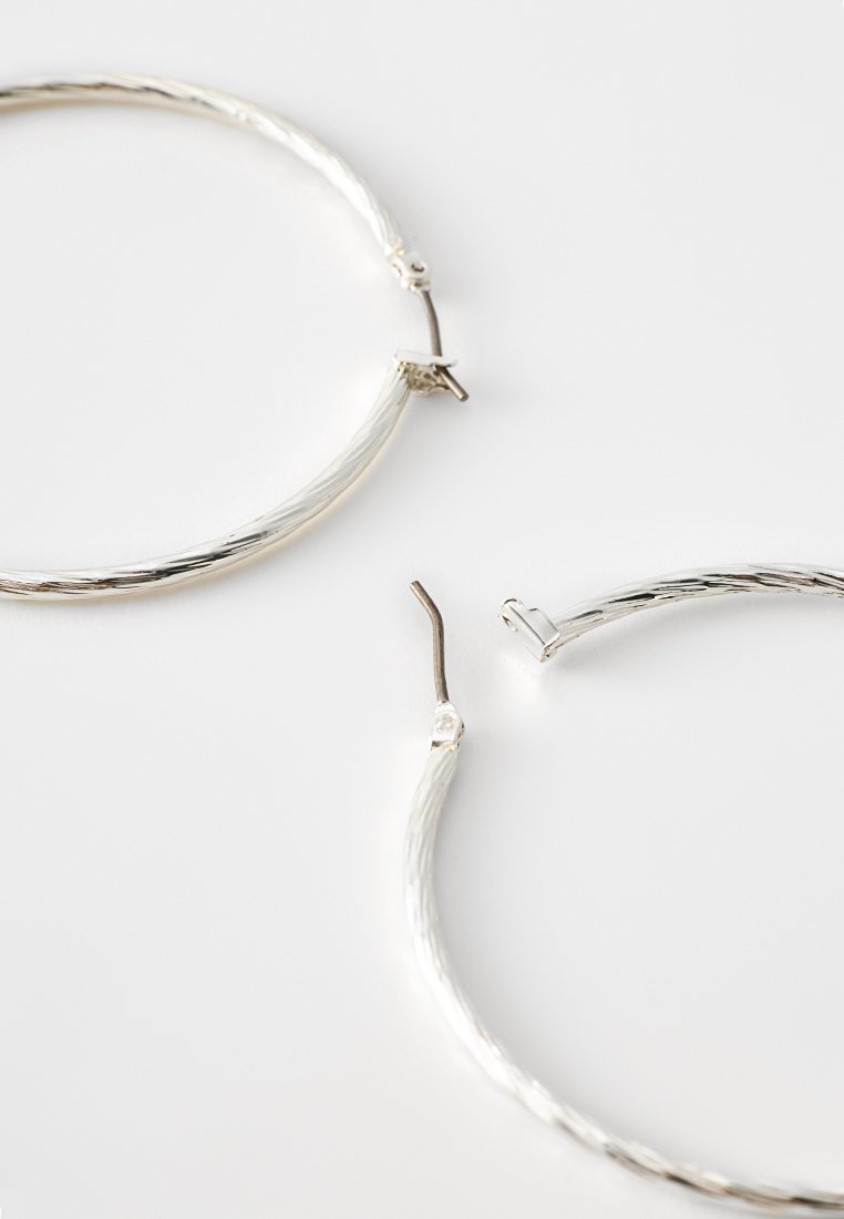 Pendientes de aro de plata con un diseño retorcido, que cuentan con un cierre de seguridad. La superficie es lisa con un acabado pulido.