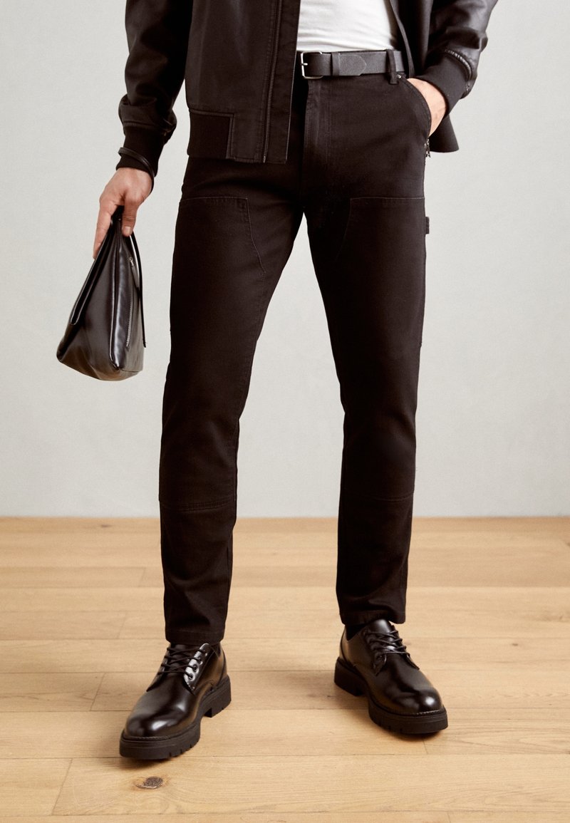 Pantalones vaqueros negros de corte slim, con bolsillos laterales y una superficie texturizada, combinados con zapatos negros de cordones pulidos.