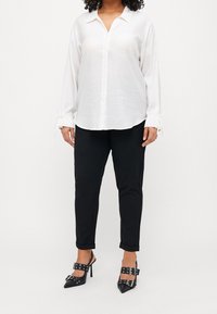 Vit, långärmad skjorta med knappar och bindningar vid ärmsluten, i kombination med svarta byxor och spetsiga svarta klackar med dekorativa spännen.