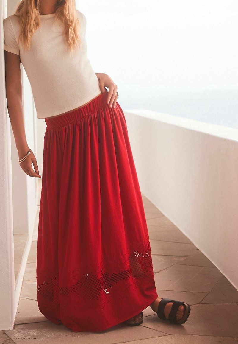Femme portant un haut blanc à manches courtes et une longue jupe rouge avec des détails ajourés, debout sur un sol carrelé contre un mur blanc.