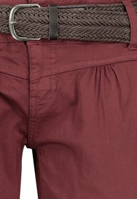 Braided brown belt with silver buckle on maroon trousers featuring a subtle gathered detail at the waistline and smooth cotton texture.