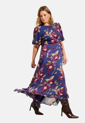 Žena v modrých kvetovaných maxi šatách s nariasenými rukávmi, kráčajúca v tmavohnedých vysokých čižmách na bielom pozadí.